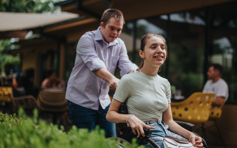 A young man with Down syndrome helps pushes a smiling girl in a wheelchair outside