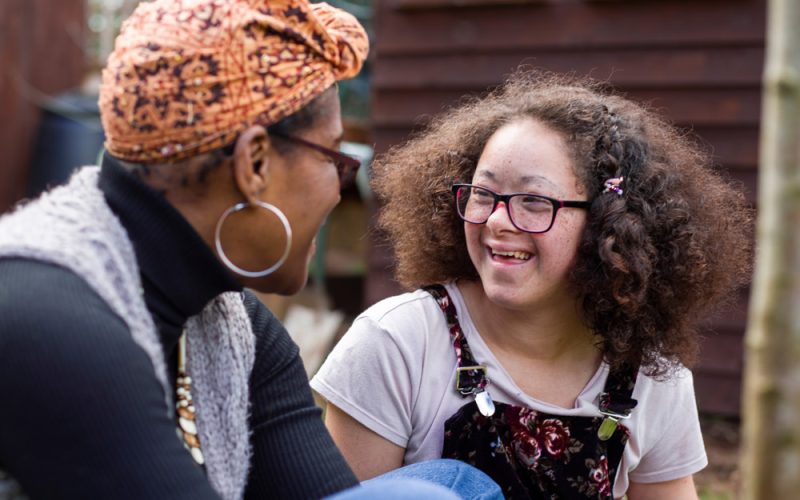 A caregiver and a young girl with curly hair and glasses sitting outside.