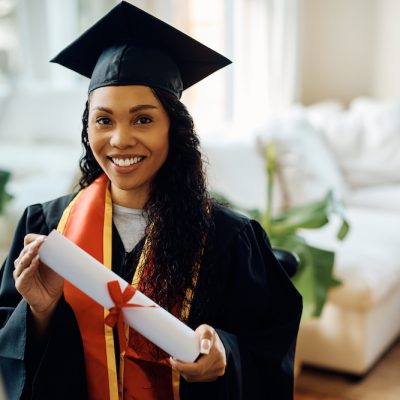 Young black woman in graduation gown holds university certificate and looking at camera.
