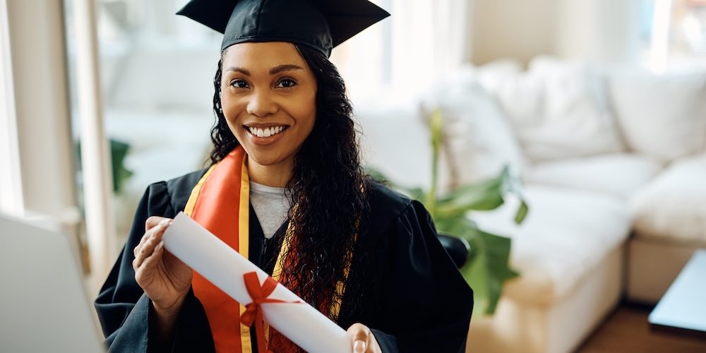 Young black woman in graduation gown holds university certificate and looking at camera.